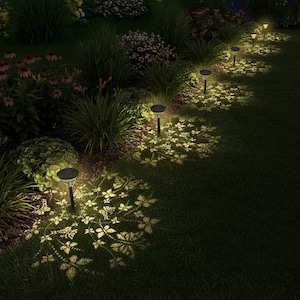 May include: A nighttime garden scene illuminated by solar-powered pathway lights. The lights cast a decorative leaf pattern onto the grass. The scene includes various plants and flowers, creating a serene and inviting outdoor space.