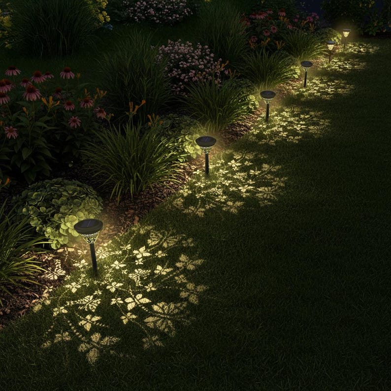 May include: A nighttime garden scene illuminated by solar-powered pathway lights. The lights cast a decorative leaf pattern onto the grass. The scene includes various plants and flowers, creating a serene and inviting outdoor space.