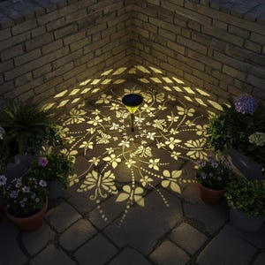 May include: A solar-powered garden light casts a butterfly and floral pattern on a brick patio. The light is surrounded by potted plants and is set against a brick wall. The light's glow creates a decorative ambiance.