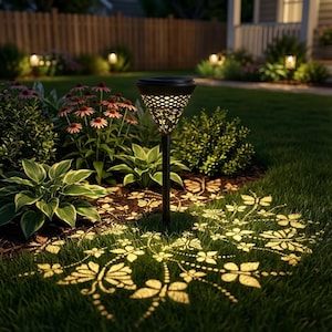 May include: A black solar-powered garden light with a decorative, patterned shade. The light casts a butterfly and floral design onto the surrounding grass. The garden is surrounded by plants and flowers, with a wooden fence in the background.