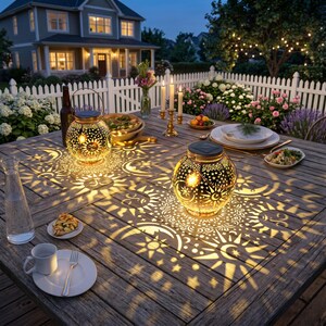 May include: Two spherical metal solar lanterns with intricate cutouts of suns, moons, and stars sit on a wooden outdoor table. The lanterns emit a warm glow, casting detailed celestial patterns onto the surface of the table in a dark setting.