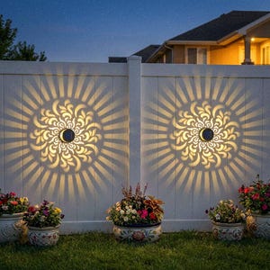 May include: White fence with two solar lights casting a decorative floral pattern. The lights are mounted on the fence and create a sunburst effect. Colorful potted flowers sit in front of the fence on a green lawn.