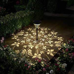 May include: A solar-powered garden light casts a butterfly and floral pattern on a stone patio. The black metal light has a decorative top and a central pole. The light is surrounded by green bushes and pink and white flowers.