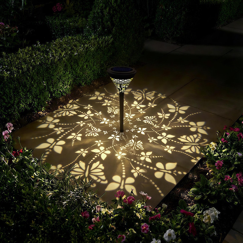 May include: A solar-powered garden light casts a butterfly and floral pattern on a stone patio. The black metal light has a decorative top and a central pole. The light is surrounded by green bushes and pink and white flowers.