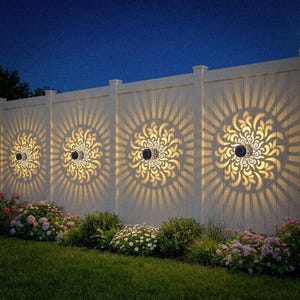 May include: Outdoor solar lights mounted on a white fence, casting decorative floral patterns. The lights are surrounded by lush greenery and flowers, creating a warm ambiance against the night sky. The lights are round and black.
