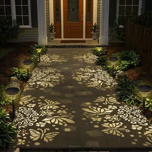 May include: Pathway illuminated by solar garden lights. The lights cast intricate butterfly and floral patterns on the concrete walkway. The scene is set in front of a house with a wooden door and surrounding greenery.