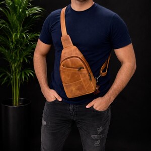 Könnte beinhalten: Eine braune Leder-Umhängetasche mit einer Reißverschlusstasche vorne und einem verstellbaren Riemen. Die Tasche wird über der Brust getragen und hat einen kleinen Anhänger. Die Tasche ist aus strukturiertem Leder.