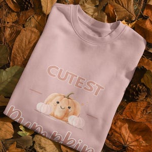 Puede incluir: Una sudadera rosa claro con las palabras "Cutest Pumpk-in" y un gráfico de calabaza sonriente. La calabaza está rodeada de dos calabazas blancas más pequeñas. La sudadera está sobre un lecho de hojas de otoño.