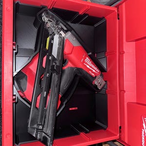 May include: A red and black Milwaukee FUEL nail gun is stored inside a red and black Packout tool case. The nail gun has a black handle and a silver and black magazine. The tool case is open, revealing the tool inside.