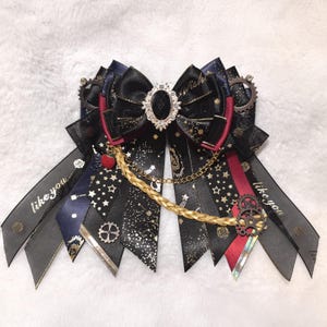 Könnte beinhalten: Dekorative schwarze Schleife mit mehreren Bändern. Sie hat einen zentralen Schmuckstein, eine goldene Kette und Akzente in Rot, Marineblau und Gold. Die Bänder tragen den Text "like you" und ein Sternenmuster.