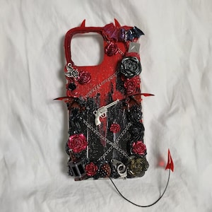 May include: A decorative phone case with a red and black design, featuring roses, a silver gun, chains, and devil horns. The case has a glittery finish and includes a small red devil tail.