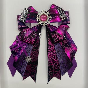 Könnte beinhalten: Dekorative Haarschleife aus lila und schwarzen Bändern mit Spinnennetz- und geometrischen Mustern. Sie ist mit einer silbernen Verzierung und einem rosa Schmuckstein in der Mitte versehen. Geeignet für Halloween oder Gothic-Themen.