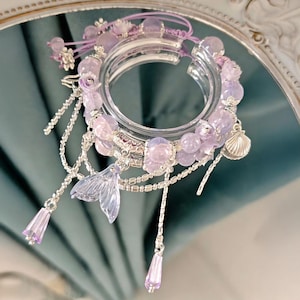 May include: A clear, circular bracelet with lavender and silver accents. The bracelet features lavender beads, dangling silver chains, a seashell charm, and a translucent mermaid tail charm. A delicate, feminine accessory.