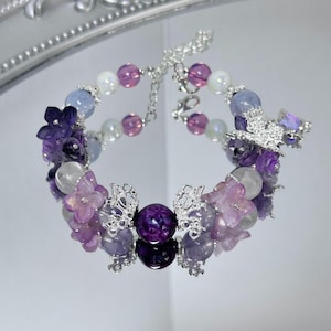 Puede incluir: Una delicada pulsera con una mezcla de cuentas moradas, lavanda y blancas. La pulsera incluye dijes de mariposa plateados y detalles de flores. Las cuentas son una mezcla de formas redondas e irregulares, creando un diseño único y elegante.