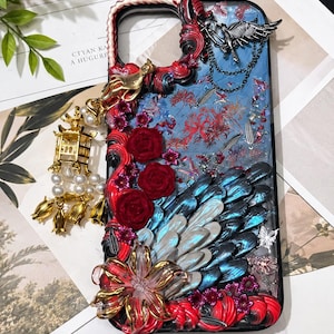 Peut inclure: Une coque de téléphone décorative avec un fond bleu à motifs floraux rouges, ornée de roses rouges en relief, d'ailes bleu et argent irisé, d'une breloque lanterne dorée avec des perles blanches et d'un ornement d'oiseau en argent.
