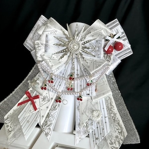 Puede incluir: Un lazo decorativo blanco y plateado con patrones y adornos intrincados. Presenta un diseño central de estrella, hileras de perlas y colgantes de cerezas. Acentuado con un lazo rojo y pequeñas cerezas rojas. Un accesorio único.