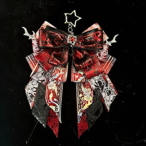 Puede incluir: Un lazo decorativo con cintas superpuestas en patrones rojos, negros y blancos. El lazo presenta un lazo central rojo con un diseño floral, un dije en forma de estrella y detalles plateados. Las cintas tienen varios patrones, incluyendo diseños florales y de reloj.