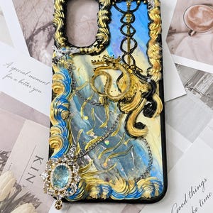 Peut inclure: Une coque de téléphone décorative avec un fond marbré bleu et jaune, ornée de volutes en relief façon crème chantilly dorées, bleues et noires. Elle comprend un dragon doré, une chaîne argentée et un grand pendentif en cristal bleu.