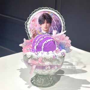 May include: A decorative dessert-themed figurine with a purple ice cream scoop on a layered glass bowl. The bowl is decorated with pink ribbons, pearls, and flowers. A circular image of a person is in the background.