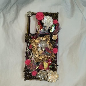 May include: A decorative phone case featuring an ornate design with a dark bronze frame. The case is adorned with pink roses, white flowers, butterflies, and gold accents. The design includes a small gold frame and various embellishments.