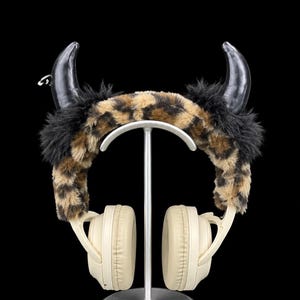 May include: Headphones featuring a leopard print faux fur headband cover with black faux fur accents and two black curved horns attached to the top. The ear cups are beige with a smooth finish, and the headband is mounted on a silver stand.
