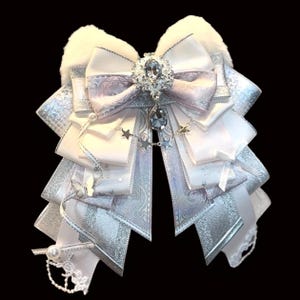 May include: A layered, decorative bow in white, silver, and pale lavender. The bow features a large, ornate rhinestone centerpiece with a dangling crystal. Silver ribbons and lace are accented with small pearls and star charms. The bow has a fluffy white top.