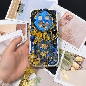 May include: A rectangular phone case featuring a deep blue and black textured background with gold and blue floral embellishments, metallic charms, and a dangling gold-toned metal ornament with intricate cutouts and small hanging chains.
