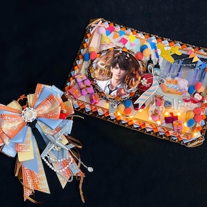 Puede incluir: Un lazo decorativo con cintas naranjas, azules y amarillas, acentuado con un dije de avión plateado y una cadena de perlas. Junto a él, un cuadro enmarcado con un retrato y decoraciones festivas, incluyendo globos y cajas de regalo.