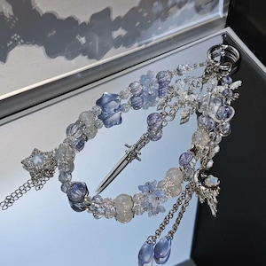 Puede incluir: Un llavero adornado con un colgante de espada en miniatura y varios abalorios. El diseño incorpora cuentas transparentes, azules y plateadas, junto con detalles de estrellas y flores. El llavero incluye cadenas colgantes y abalorios en forma de lágrima.