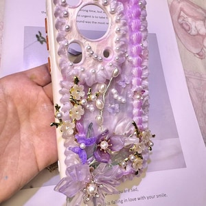 May include: A white phone case with a decorative floral design. The case features purple and white flowers, pearls, and a textured border. The text on the paper reads "wrong time, but..." and "because you never... who is falling in love with your smile."
