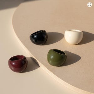 Op de afbeelding: Vier glanzende, koepelvormige ringen in zwart, wit, bordeauxrood en olijfgroen. De ringen hebben een glad, afgerond ontwerp en worden gepresenteerd op een neutraal getinte ondergrond.