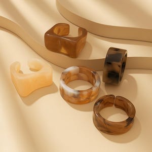 以下が含まれることがあります： 茶色、ベージュ、クリーム色など、さまざまな色合いの5つの分厚い樹脂リングのコレクション。 リングは、正方形と円形のデザインなど、さまざまな形状をしています。 リングはニュートラルな背景に表示されます。