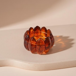 Può includere: Un braccialetto marrone a forma ovale con un motivo tartarugato. Il braccialetto è composto da più segmenti collegati da un elastico. I segmenti sono di colore ambra traslucido, creando un effetto a strati. Il braccialetto è mostrato su una superficie neutra.