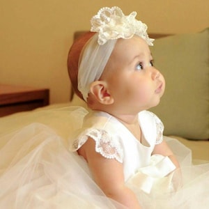 May include: A baby girl wearing a white tulle dress with a white lace headband. The headband has a large white flower on top.