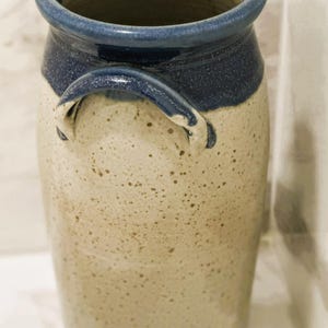 May include: A handmade ceramic vase with a speckled beige body and a dark blue rim and handle. The vase has a cylindrical shape and a wide opening. The handle is arched and attached to the side of the vase.