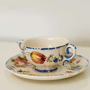 Puede incluir: Un juego de taza y platillo de color crema. La taza tiene dos asas y está decorada con un diseño floral en tonos rojos, amarillos, azules y verdes. Los bordes de ambas piezas están adornados con ribetes azules.