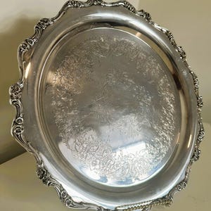 Puede incluir: Una bandeja de servir plateada y ornamentada con un detallado diseño floral en el centro. La bandeja tiene un borde festoneado con detalles decorativos. La superficie reflectante sugiere un acabado pulido, adecuado para servir alimentos o como pieza decorativa.