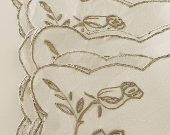May include: A stack of off-white scalloped-edge placemats. Each placemat features an embroidered floral design with a rose and leaves in a muted gold colour. The placemats are made of a textured fabric.