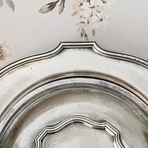 Op de afbeelding: Een zilverkleurig metalen serveerblad met een geschulpte rand en een verhoogd, meerlaags randontwerp. Het oppervlak van het blad heeft een gepolijste, reflecterende afwerking met subtiele tekenen van slijtage en aanslag langs de rand.