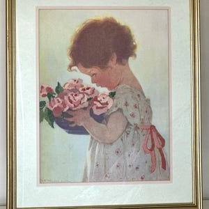 May include: A framed needlepoint art piece featuring a child with curly brown hair wearing a white floral dress with a pink ribbon, holding a blue bowl filled with pink roses. The artwork is encased in a gold-toned metal frame.