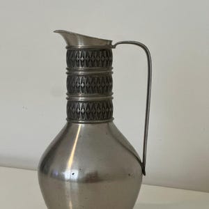 May include: A silver-colored pitcher with a rounded body and a decorative band of diamond-shaped patterns. The pitcher has a spout and a curved handle. The metal pitcher is likely vintage and suitable for serving beverages.