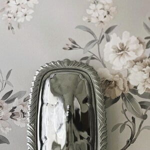 May include: A rectangular silver butter dish with a polished, reflective surface and a domed lid. The lid features a decorative, scalloped edge with intricate fluted detailing and a small, central handle for lifting the cover from the base.
