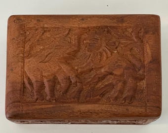 May include: A rectangular wooden box with a carved relief depicting elephants and foliage. The box is a warm brown color and appears to be made of hardwood. The carving is detailed and shows the animals and plants in a naturalistic style.