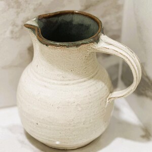 May include: Cream-colored ceramic pitcher with a rounded body and a ribbed handle. The rim and interior have a dark green glaze with a brown edge. The pitcher has a spout for easy pouring and is suitable for serving beverages.