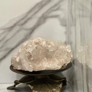 Puede incluir: Un racimo de cristales de cuarzo rosa pálido y translúcido sobre un soporte circular de metal en tono bronce oscuro. El soporte presenta una base decorativa con forma de hojas estilizadas y superpuestas con un acabado texturizado.
