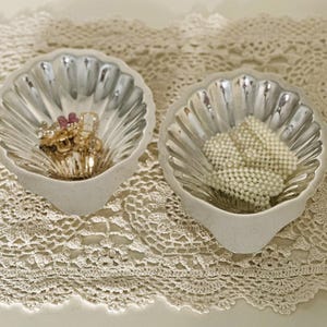 May include: Two seashell-shaped trinket dishes. One dish holds gold-toned jewelry with pink accents, the other holds a collection of white pearl beads. The dishes are white with a silver interior and sit on a cream-colored lace doily.