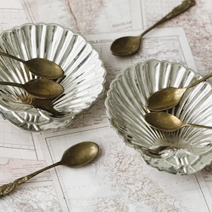 May include: Two silver-toned metal dishes shaped like scallop shells, each featuring a fluted design. Five small, tarnished brass-colored spoons with ornate handles are arranged inside and around the two shell-shaped dishes.
