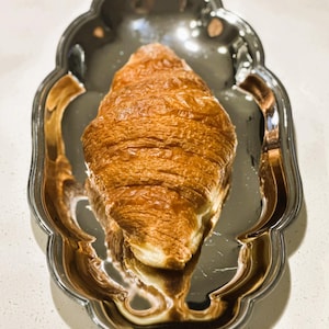 May include: A golden-brown croissant rests on a silver, scalloped-edge serving tray. The pastry's flaky layers are visible, and the tray reflects the surrounding light. The image is a close-up, highlighting the textures and details of the baked good.