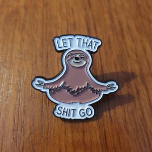 Könnte beinhalten: Anstecknadel mit einem braunen Faultier in Yoga-Pose und dem Text "LET THAT SHIT GO" in weißen Buchstaben auf schwarzem Hintergrund. Das Faultier lächelt und ist von einem weißen Rand umgeben. Die Nadel liegt auf einer Holzoberfläche.