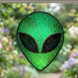 May include: A green stained glass alien head with black eyes and a textured surface hangs in front of a window. The alien is suspended by a chain. The background shows a blurred garden scene with flowers.
